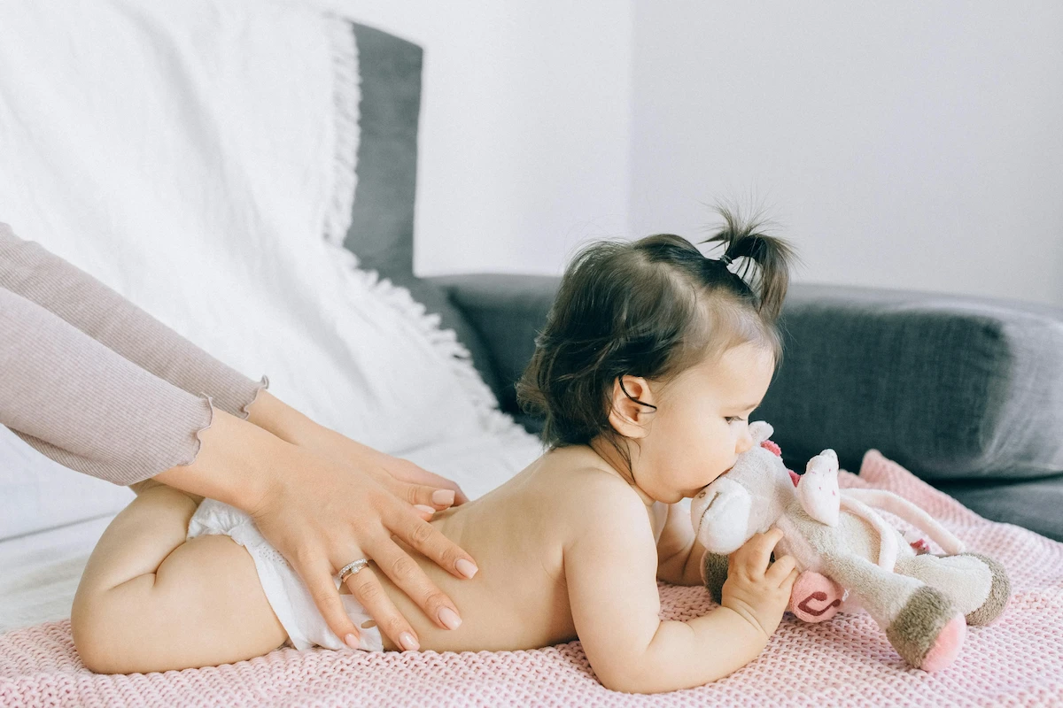 A baby lying on its stomach on a pink blanket, holding a stuffed animal, with an adult's hands gently on its back.