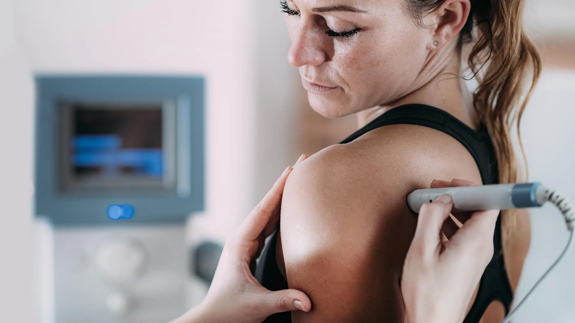 A woman receives shoulder treatment from a handheld device, with a medical machine in the blurred background.