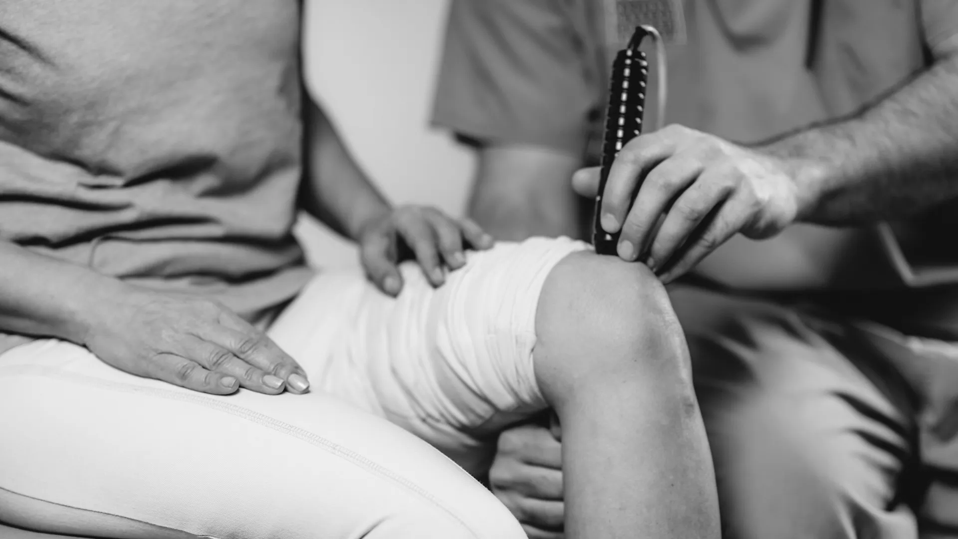 Black and white image of a therapist using a handheld device on a patient's bandaged knee.