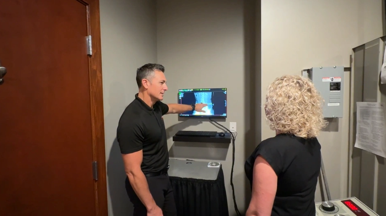 A man in a black shirt points at a medical scan on a computer screen while a woman with curly blonde hair looks on.
