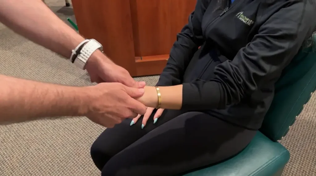 A person in a black Healthfirst Chiropractic top sits in a green chair while a practitioner gently holds their wrist, revealing a gold bracelet and light blue nails.