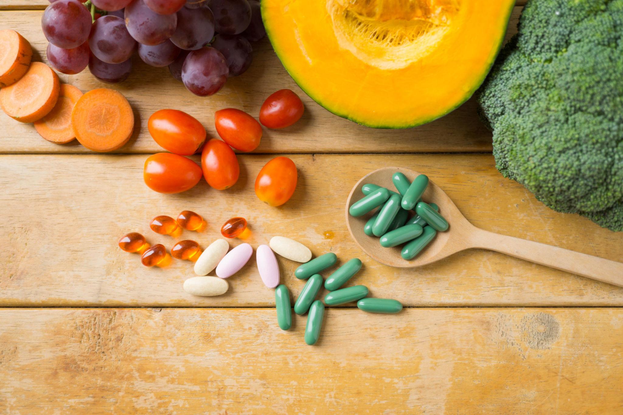 Overhead view of grapes, carrots, tomatoes, pumpkin, and broccoli with various colored pills and capsules on a wooden surface.
