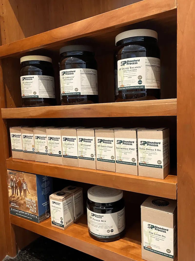 A wooden shelf unit displaying various Standard Process dietary supplements, including large jars of protein powders and detox blends, and smaller boxes of vitamins, minerals, and oils.