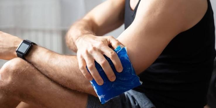 A person applies a blue gel ice pack to their elbow, wearing a black tank top and a smartwatch.